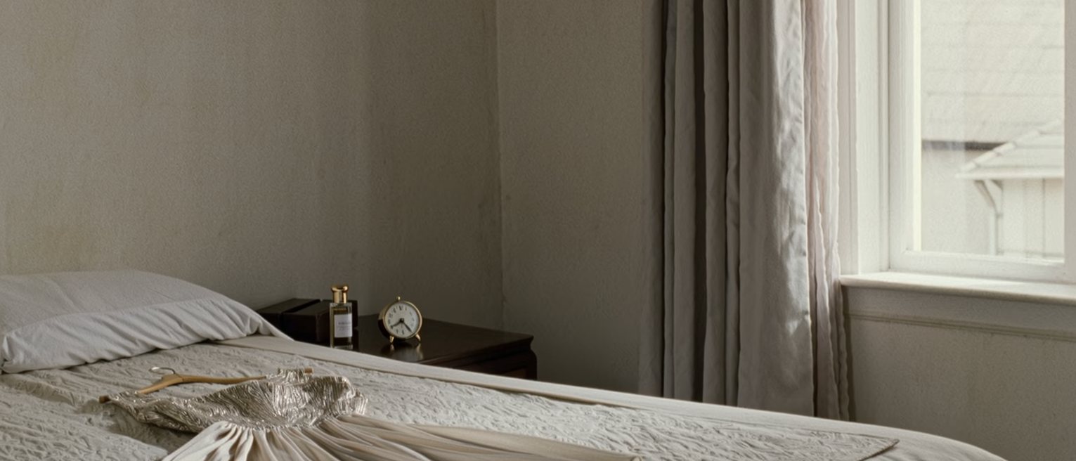 a bedroom seen close with the bed made tight, a dress laid out on it and a bottle of scent on the bedside table beside a small clock, the curtains open behind