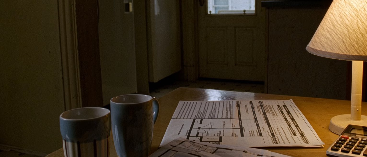 a flat's kitchen table seen close with a housing board application form beside two mugs, a calculator, a folded floor plan and a tin of biscuits, the rice cooker and the shelves close behind and the window onto the block opposite soft beyond