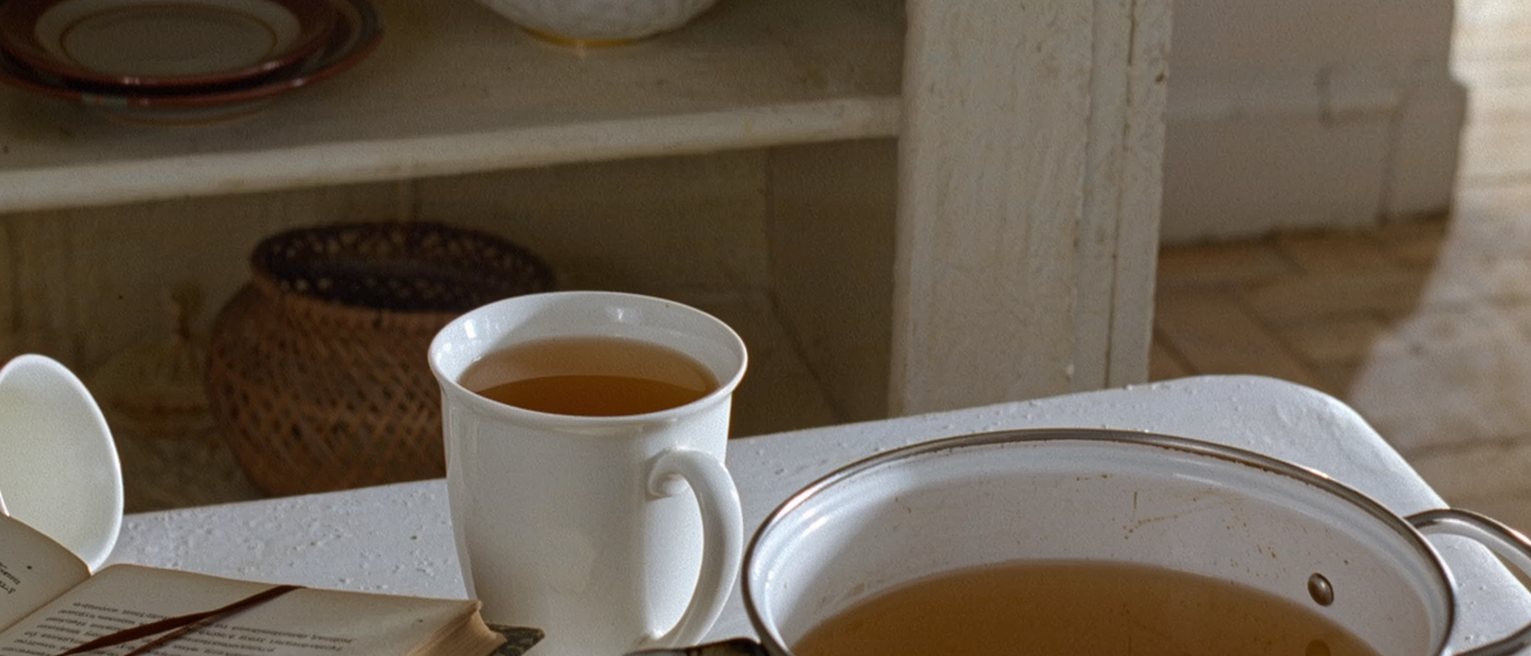 a kitchen table seen close with a pot of tea and one cup poured, the second cup upturned and waiting, a book open with a ribbon marking the page and the shelves soft behind