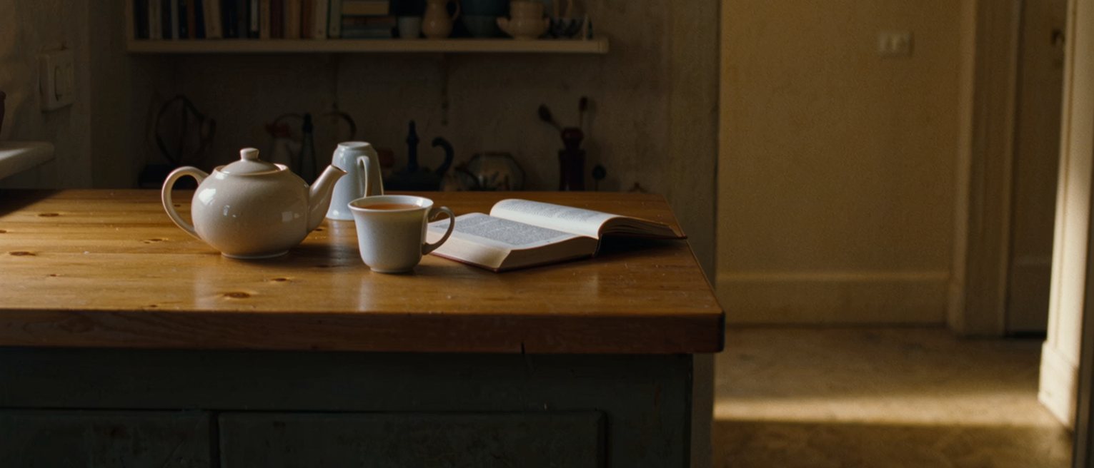 a kitchen table seen close with a pot of tea and one cup poured, the second cup upturned and waiting, a book open with a ribbon marking the page and the shelves soft behind
