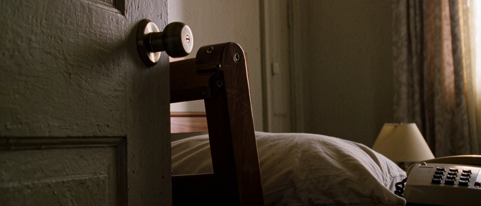 a bedroom seen close with a door bolt drawn and a chair wedged under the handle, a phone on the pillow and a lamp, the curtains drawn behind