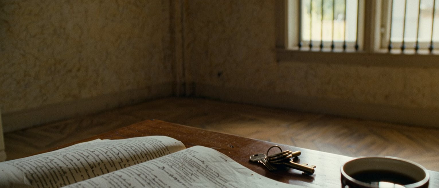 a kitchen table seen close with a printed constitution open at its first pages, a cup of rooibos, a set of keys and the breadboard with its loaf, the dresser with its plates close behind and the barred window soft beyond