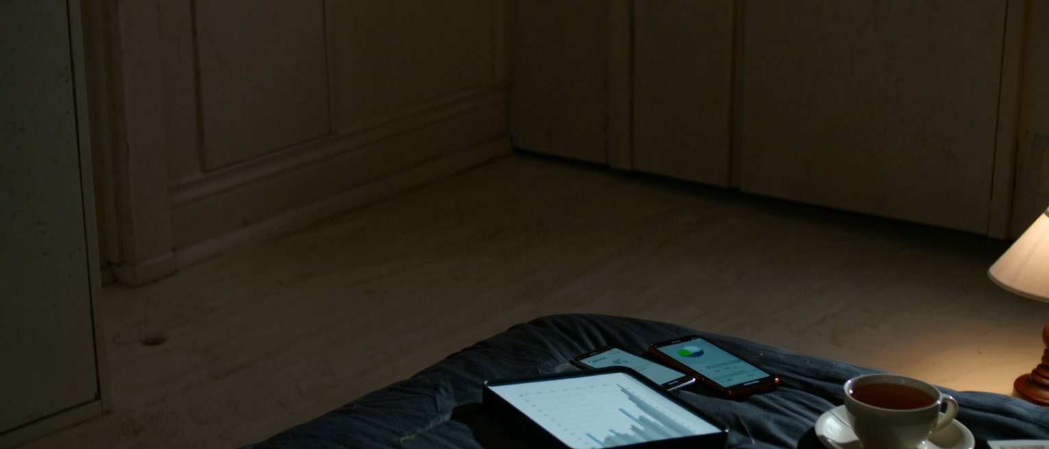 a bedroom seen close with two phones charging, a printed report with a graph folded open on the bed, a cup of tea and a paperback, the lamp close behind