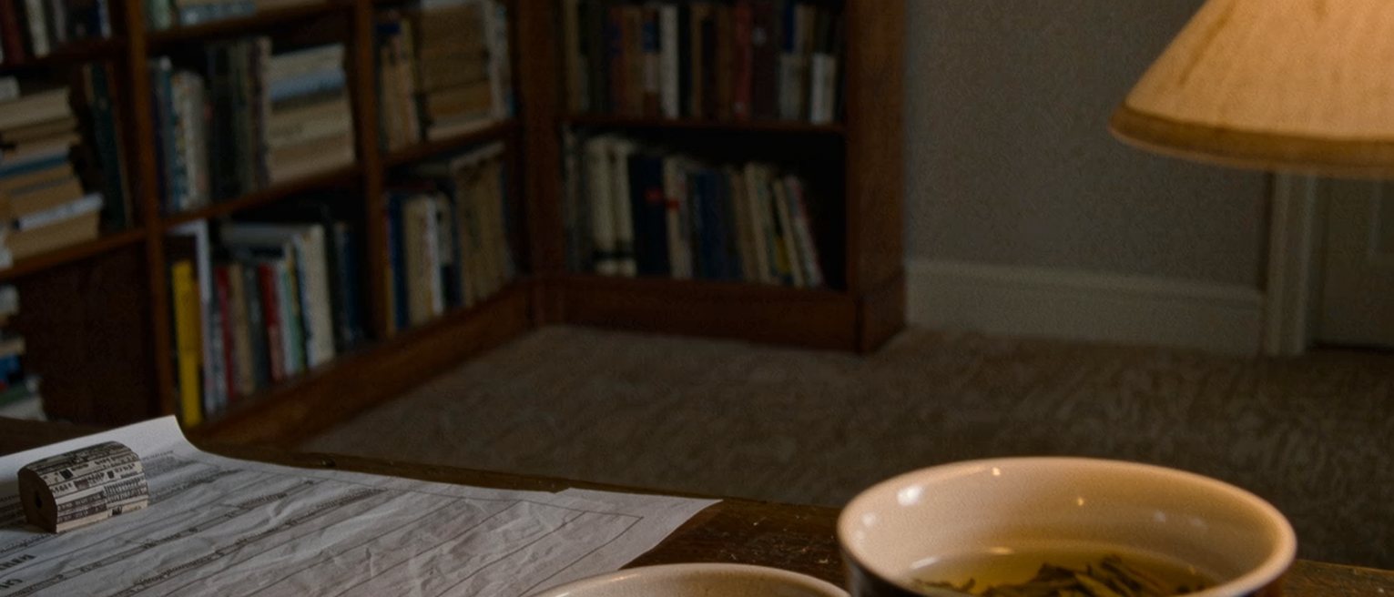 a desk seen close with a ballot paper stub beside a ruling printed and stapled, two rings in a dish and a cup of oolong, the bookshelves soft behind
