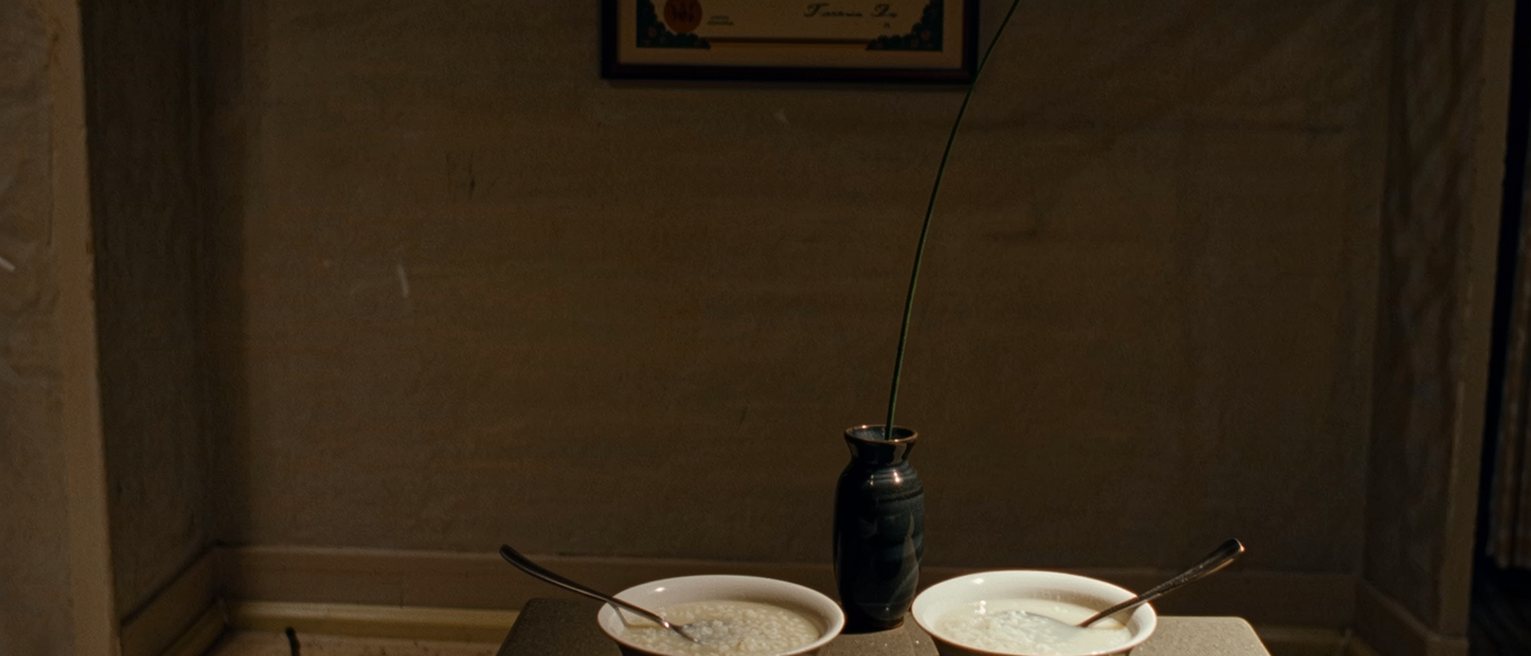 a kitchen seen close with two bowls of congee, two spoons and a small vase with a single stem, a framed certificate on the wall behind