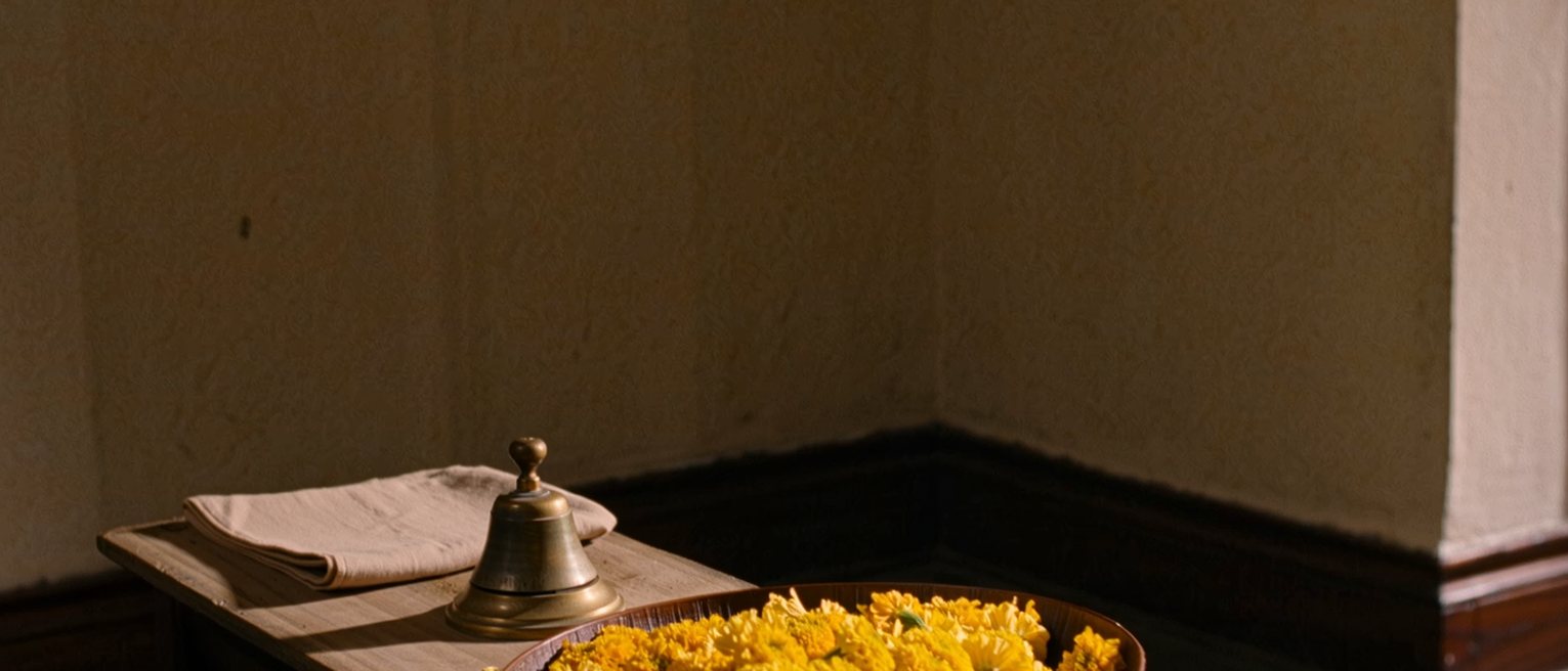 a low altar shelf seen close in a private room with a single brass bell, a folded cloth and a bowl of marigold petals, the plain wall soft behind