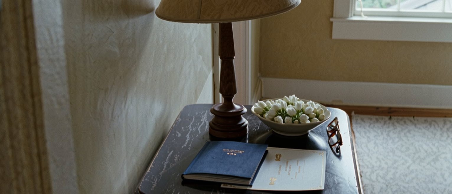 a hall table seen close with a blue household registration book closed beside a folded marriage certificate, a pair of reading glasses and a bowl of jasmine, the lamp close behind