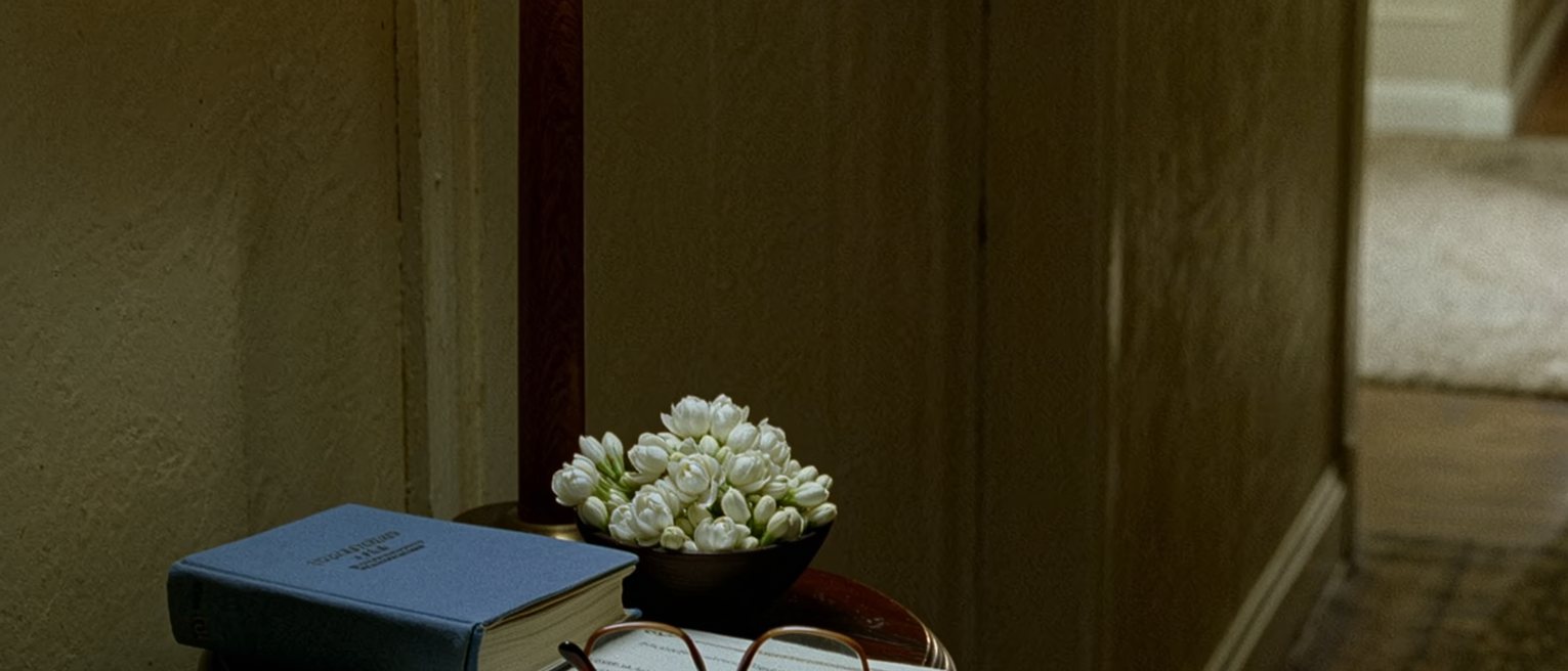 a hall table seen close with a blue household registration book closed beside a folded marriage certificate, a pair of reading glasses and a bowl of jasmine, the lamp close behind