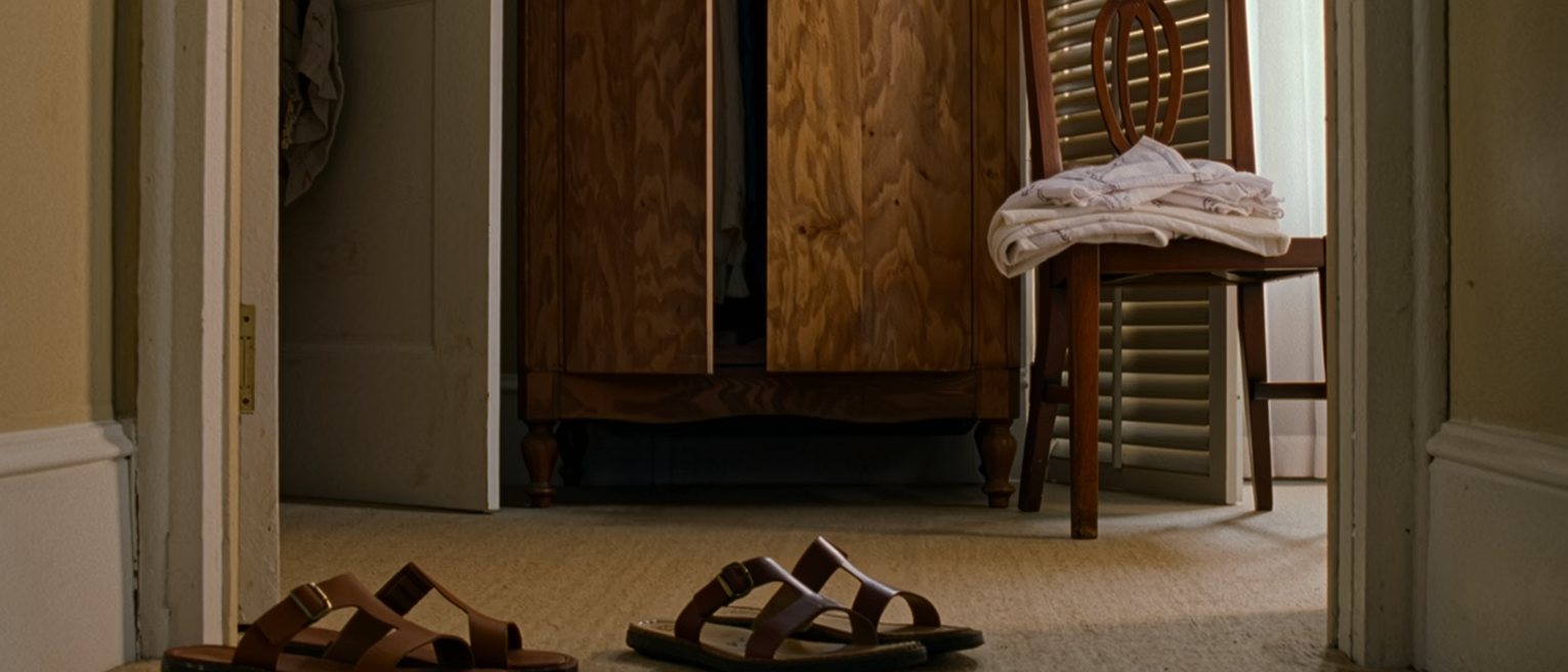 a bedroom seen close with two pairs of sandals by the door, a folded sarong on the chair and a wardrobe shared down the middle, the shutters open behind