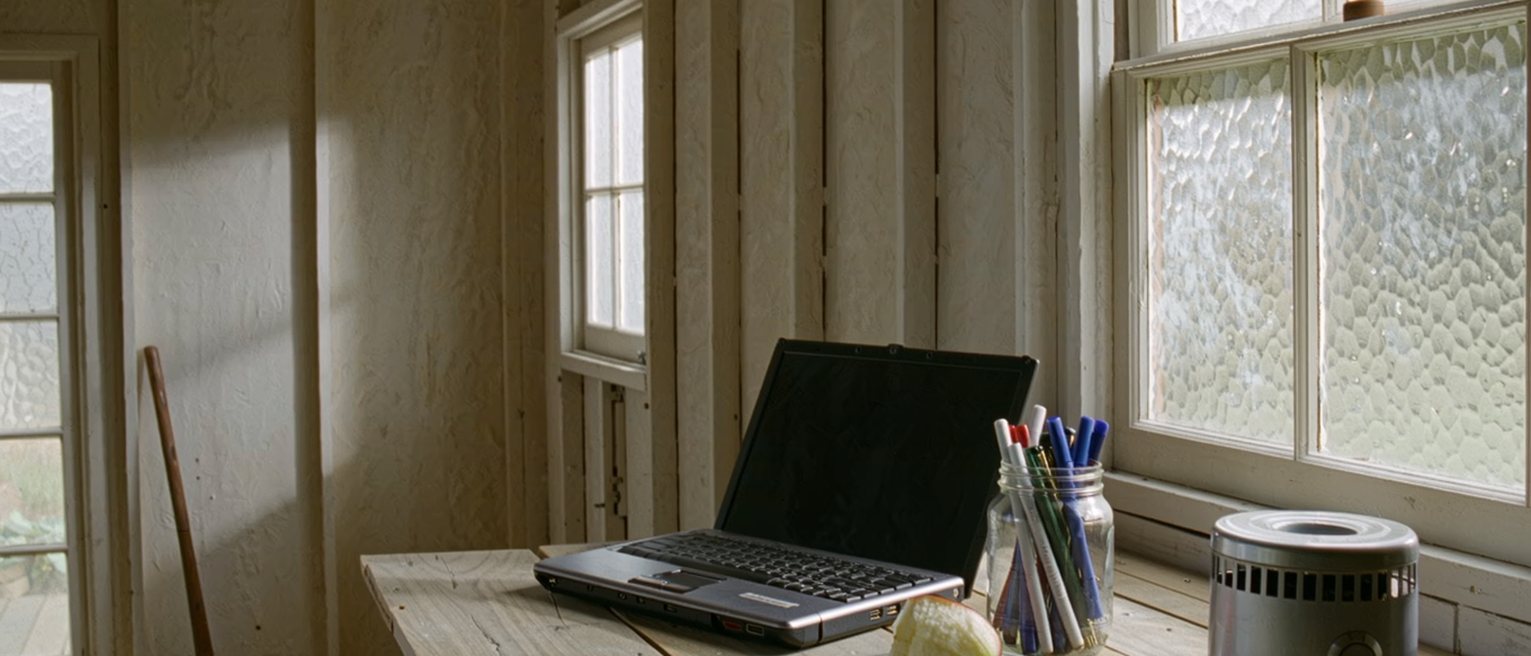 a garden shed seen close with a laptop shut on a plank desk, a jar of pens, an electric heater and an apple core on a saucer, the frosted window soft behind