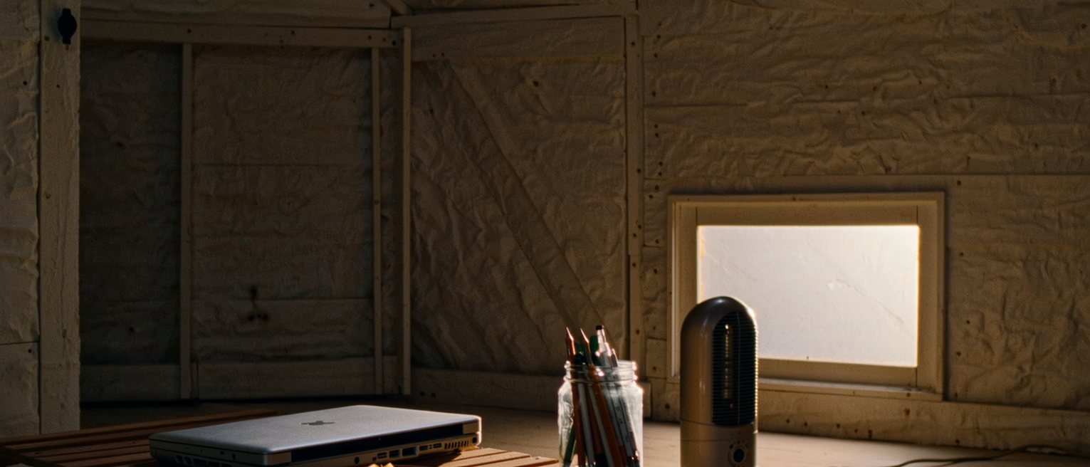 a garden shed seen close with a laptop shut on a plank desk, a jar of pens, an electric heater and an apple core on a saucer, the frosted window soft behind