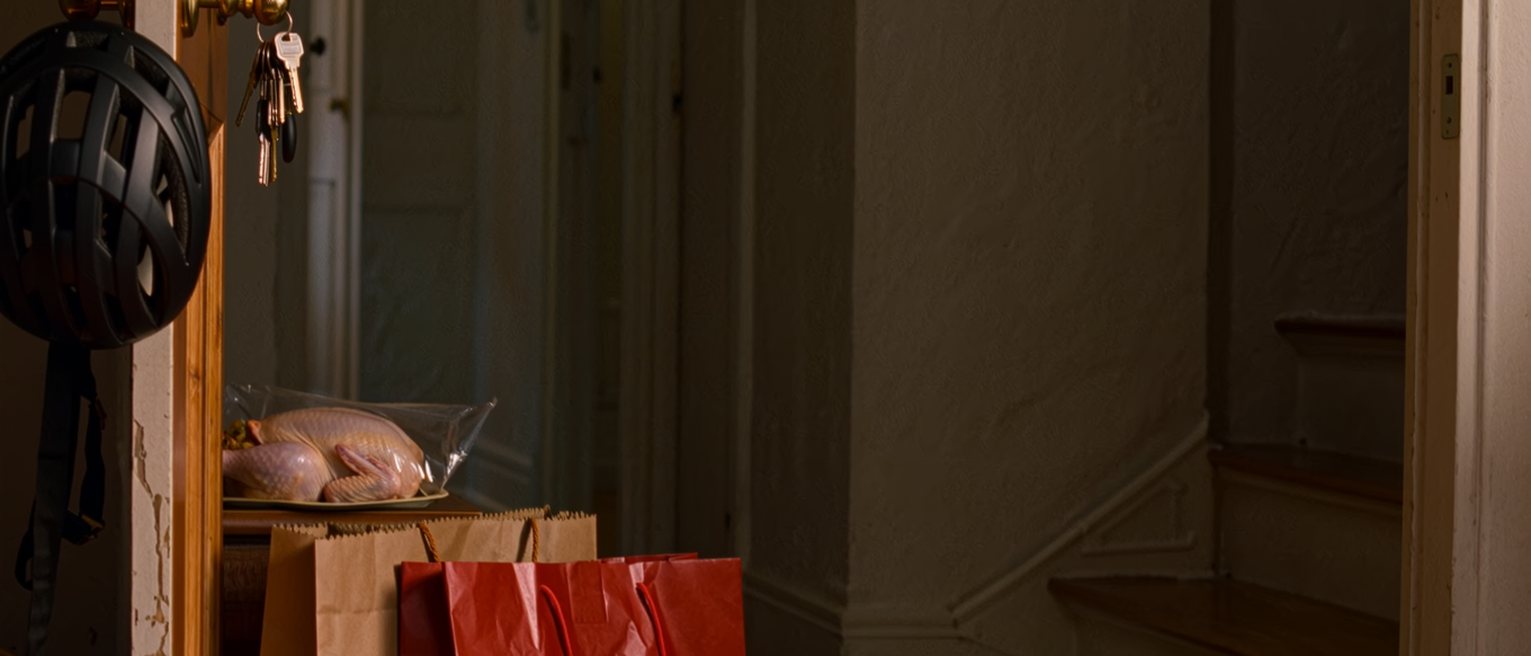 a hall seen close with a bicycle helmet on the hook, two shopping bags set down on the floor, a bunch of keys still in the door and a raw chicken sealed in its plastic tray on the console, the stairwell soft behind