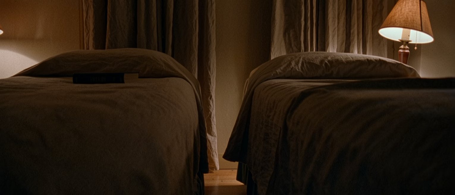 a bedroom seen close with two single beds pushed together and a gap opened between them, a lamp on each side and one book on the near pillow, the curtains soft behind