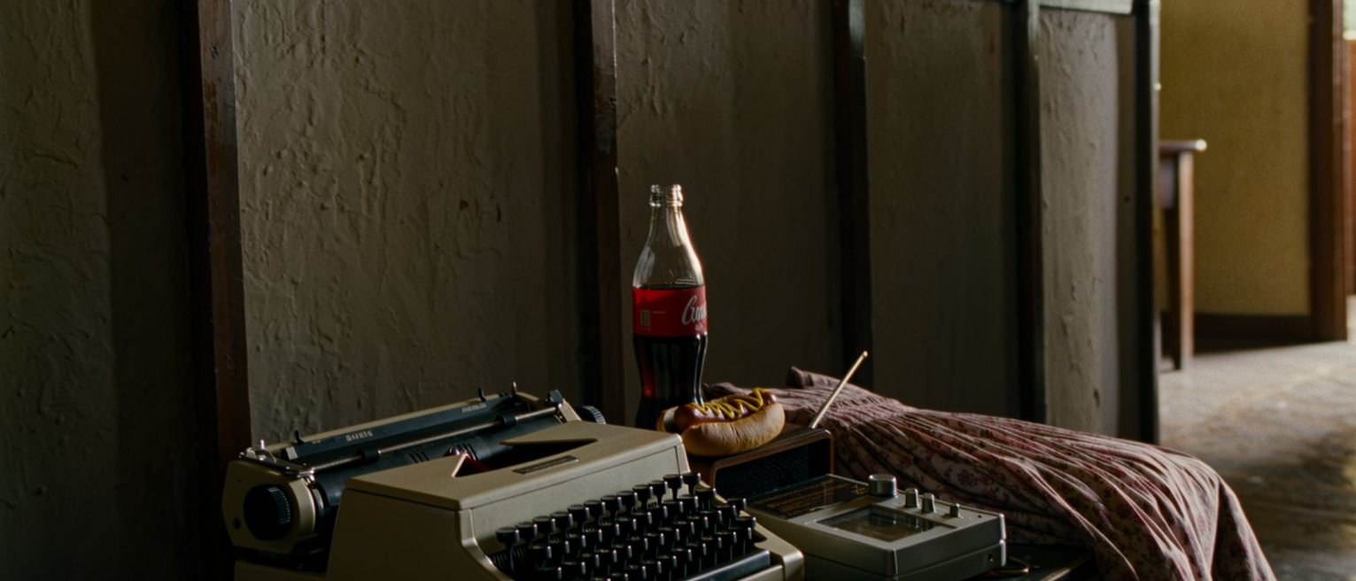 a rented room seen close with a typewriter on a small table, a bottle of cola half drunk, a radio playing beside a folded dress and a hot dog wrapper, the partition wall soft behind