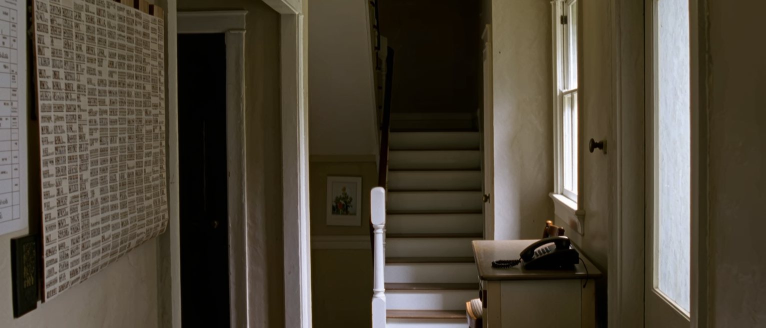 a hall seen close with a wall calendar crowded with other names, a shopping list pinned beside it, a landline phone on the console and a chair beneath, the stairs close behind