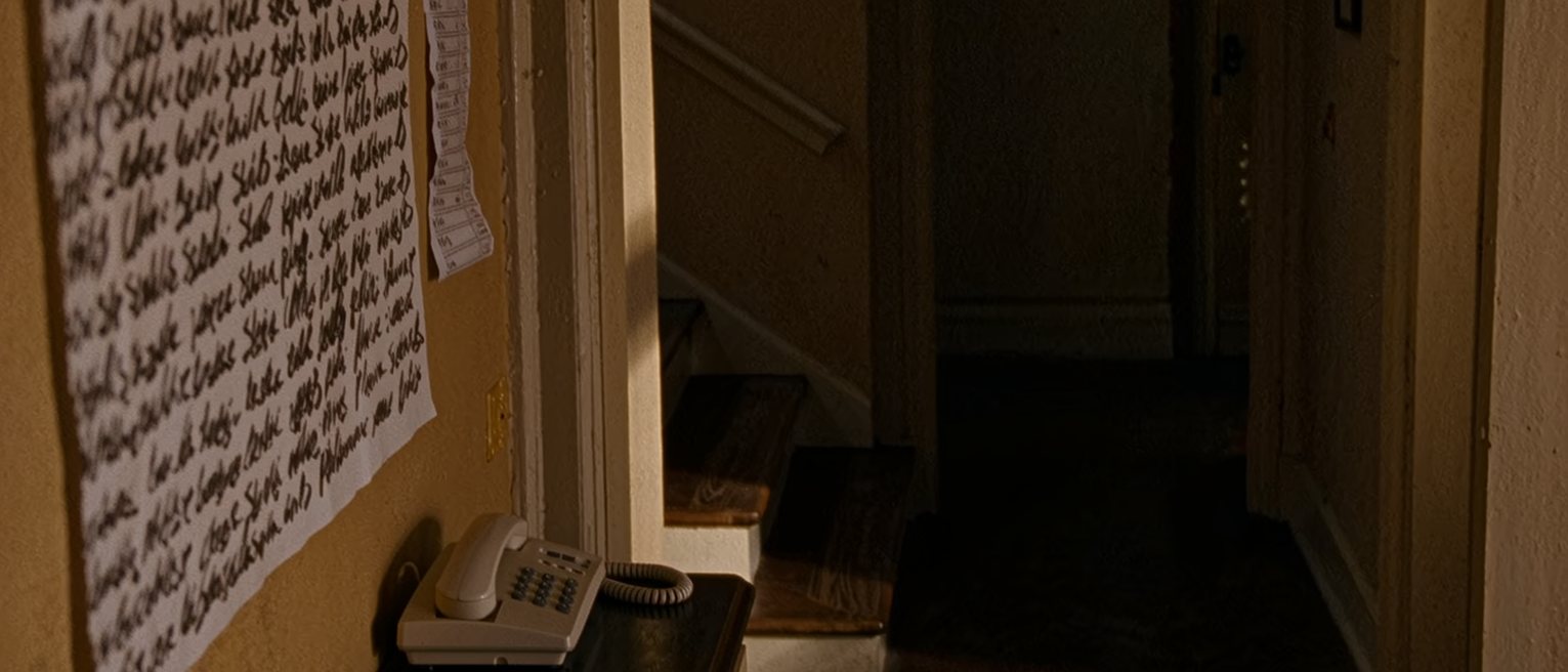 a hall seen close with a wall calendar crowded with other names, a shopping list pinned beside it, a landline phone on the console and a chair beneath, the stairs close behind