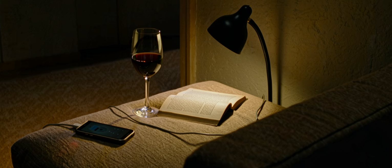 a sofa seen close with a phone face up with its screen dark on the cushion, a glass of wine and a paperback, a charger cable trailing to the wall, the lamp close behind