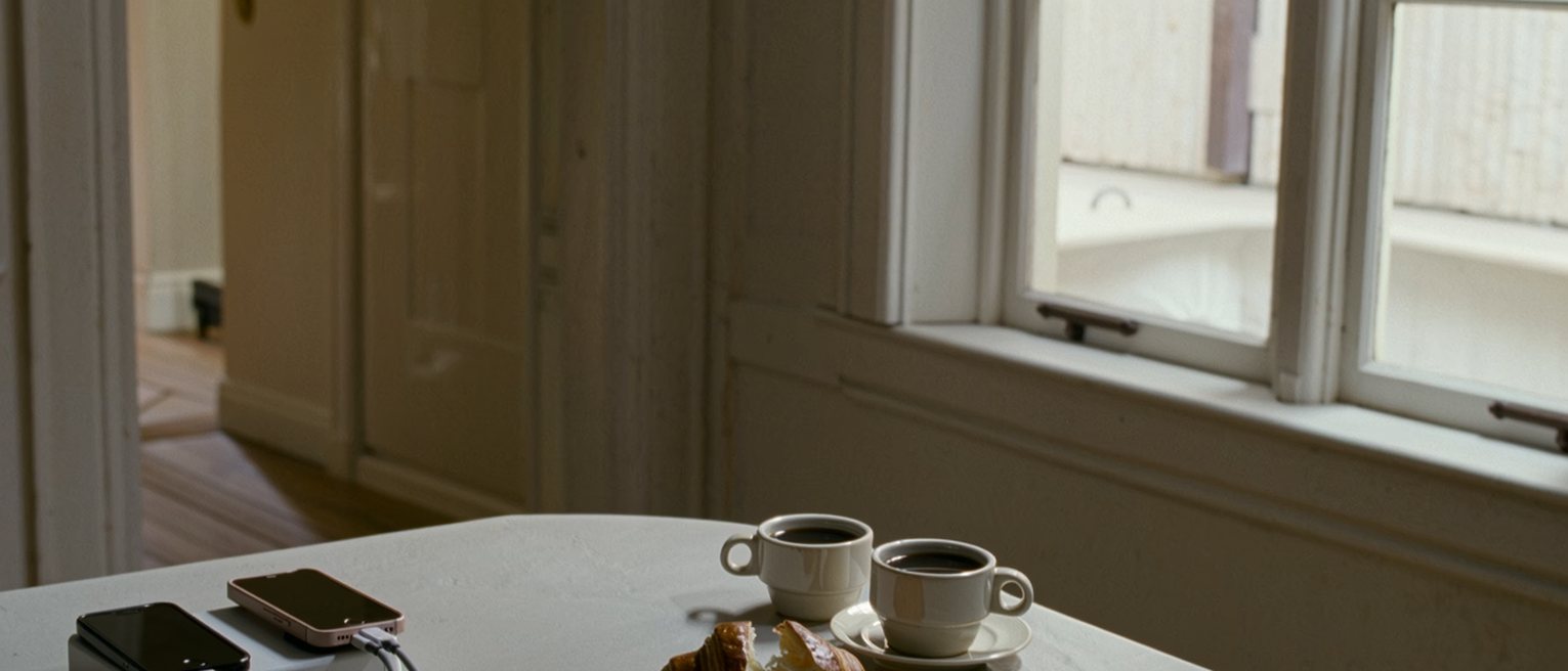 a kitchen table seen close with two phones charging from one adaptor, two coffees and a croissant torn in half, a printed boarding pass folded, the window soft behind