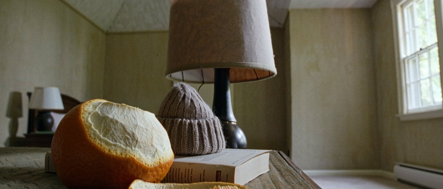 a bedside table seen close with an orange peeled in one long spiral, a small knitted hat and a paperback, the lamp close behind