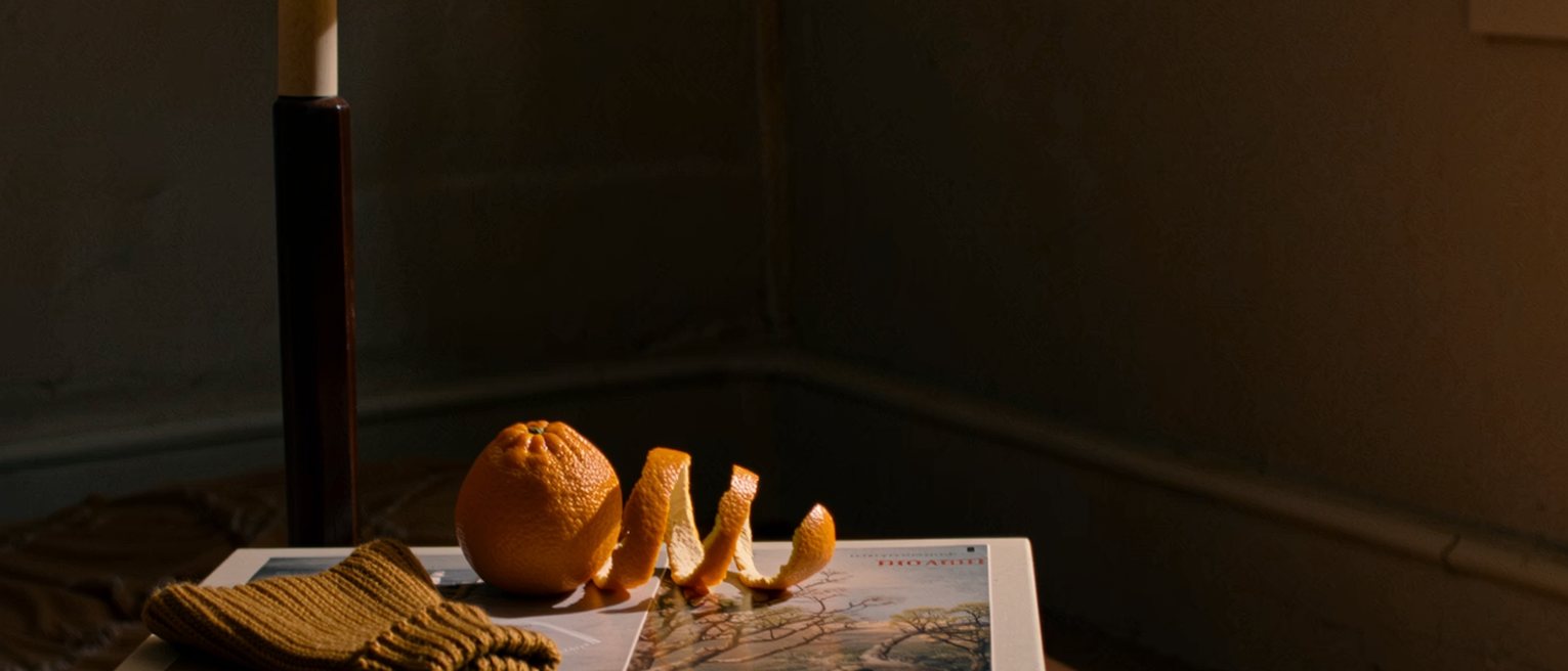 a bedside table seen close with an orange peeled in one long spiral, a small knitted hat and a paperback, the lamp close behind