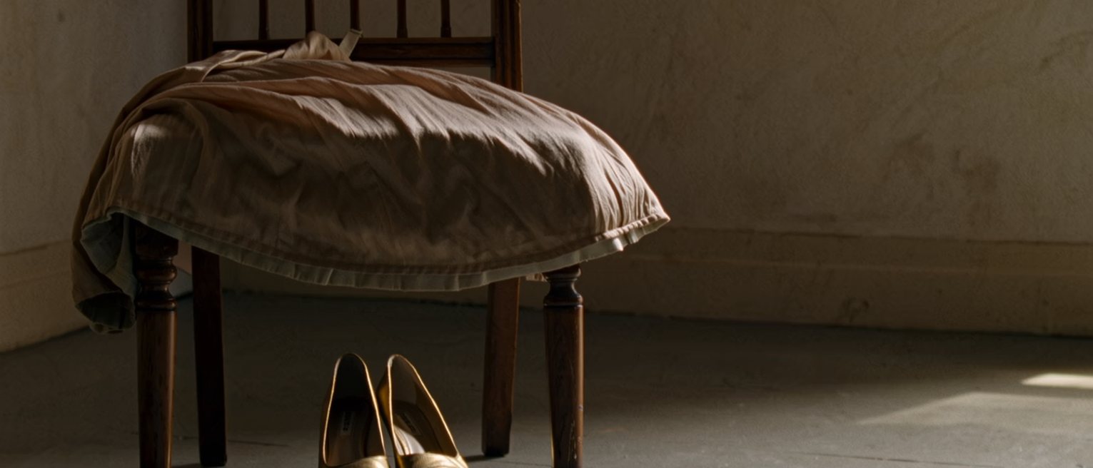 a bedroom with a worn silk dress laid over a chair and a pair of gold shoes beneath it, the made bed and a dressing table close behind