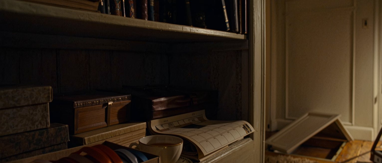 a study seen close with a shelf of objects with gaps where things stood, a box of ribbons sealed shut, a calendar with its pages torn out and a cup of tea, the desk lamp and the bookshelves close behind