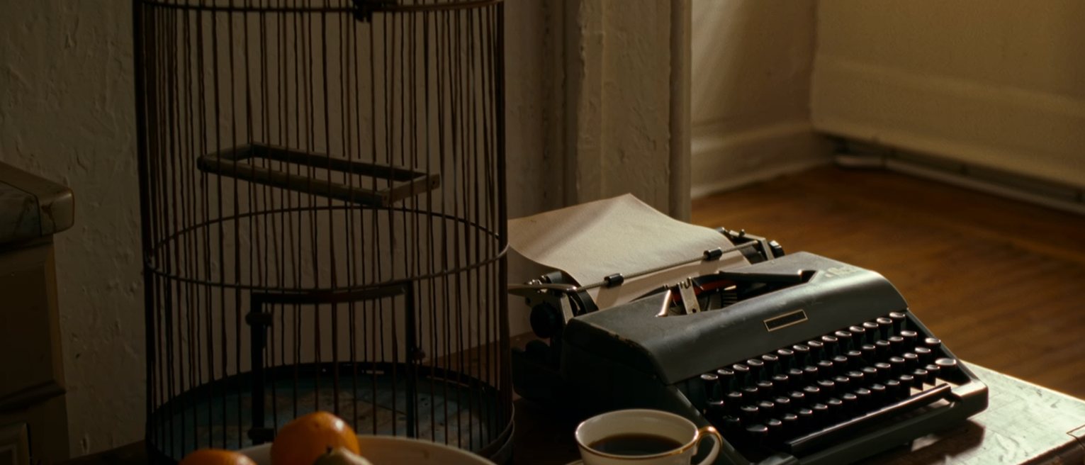a kitchen table seen close with a bowl of fruit and one kind missing, a bird cage empty with its door open, a typewriter with a blank page and a cup of coffee, the window soft behind
