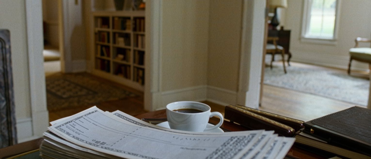 a desk seen close with a film camera, a stack of unpaid bills fanned beside a cheque book and a pawn ticket, a cup of coffee and the bookshelves soft behind