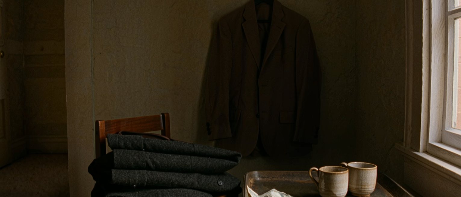 a bedroom seen close with a good coat folded over the chair, a jacket on the hook beside it, a tray with two cups and a bill tucked under one, the window soft behind