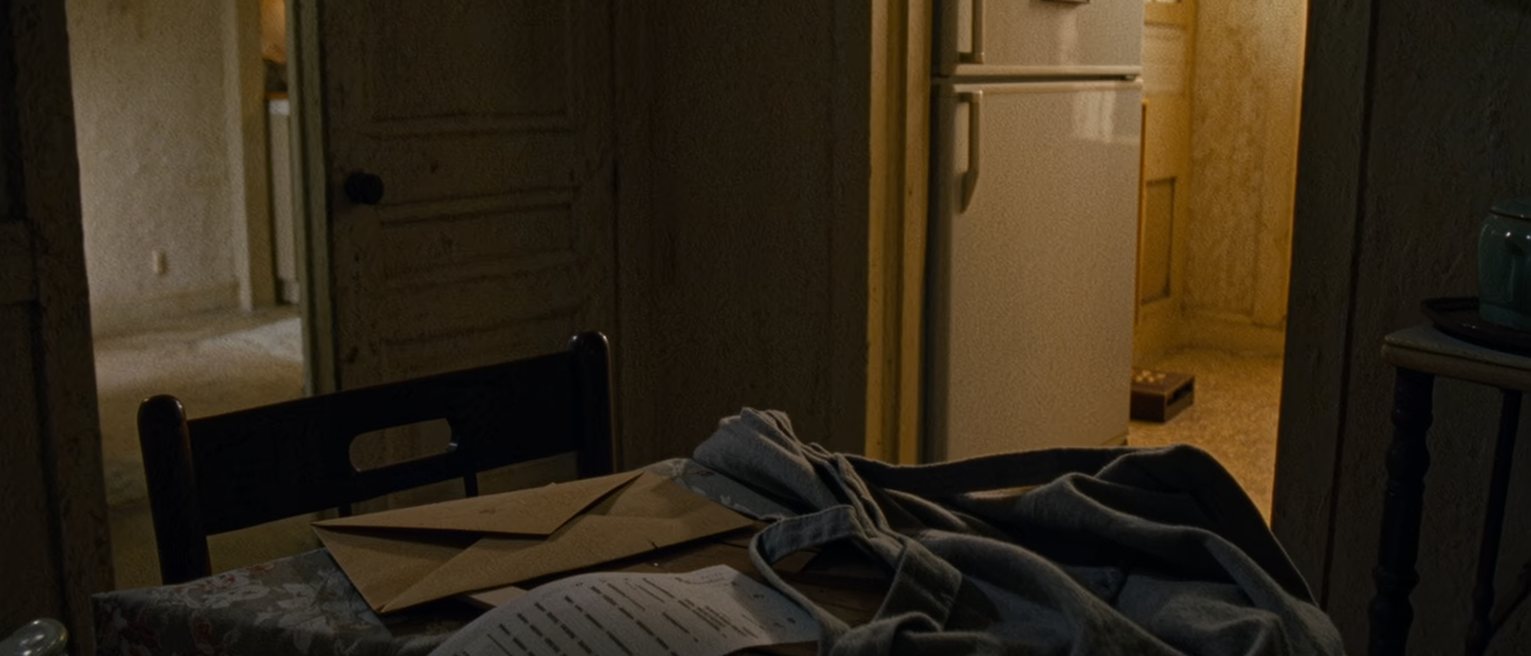 a kitchen seen close with a pay envelope on the table beside a remittance receipt and a phone face up, a folded apron on the chair and a small framed drawing on the fridge, the window soft behind