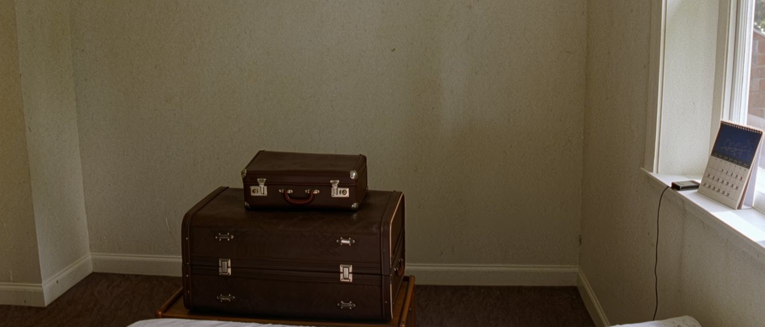 a bedroom seen close with a single bed in a small room, a suitcase on top of the wardrobe, a calendar with a date circled in a far month and a phone charging on the sill, the window soft behind