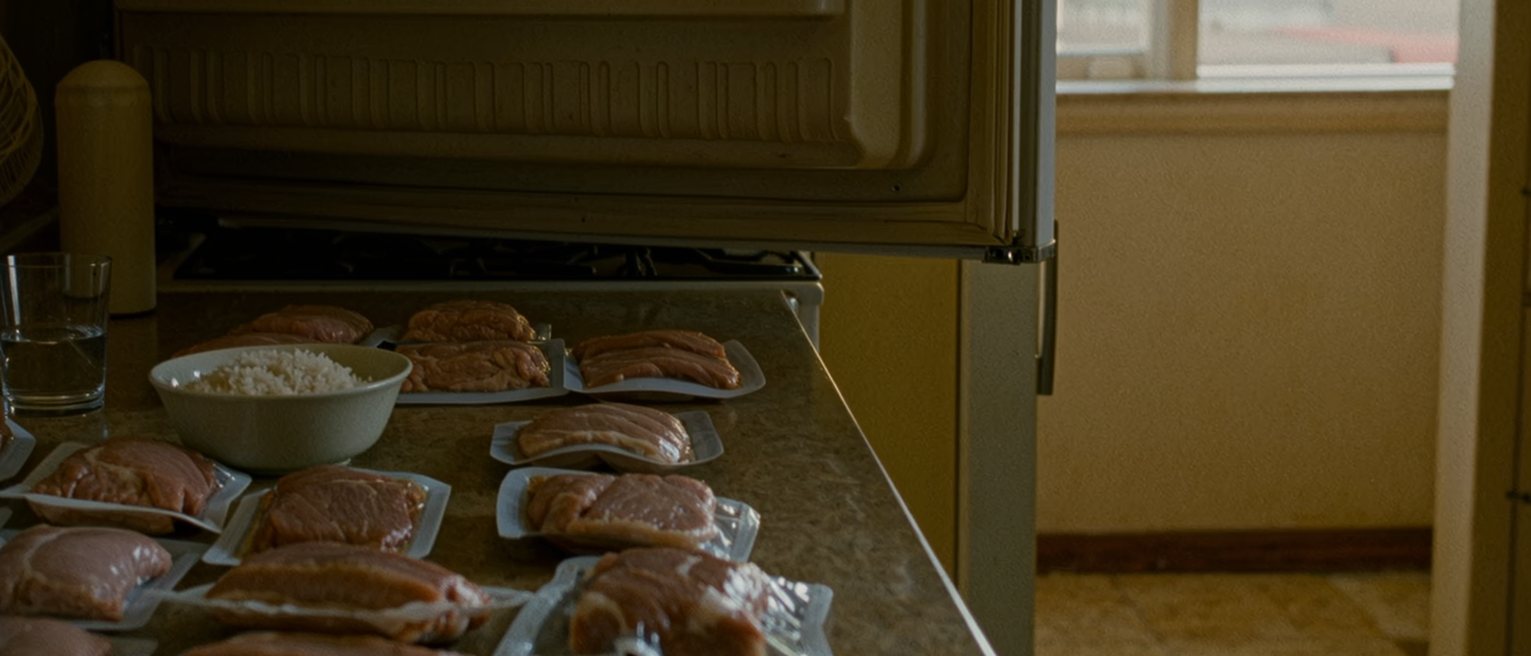a kitchen counter seen close with the fridge open beside it and the meat cleared out onto the counter in its packets, a bowl of plain rice and a glass of water at one place, the cooker and the shelves close behind and the window soft beyond