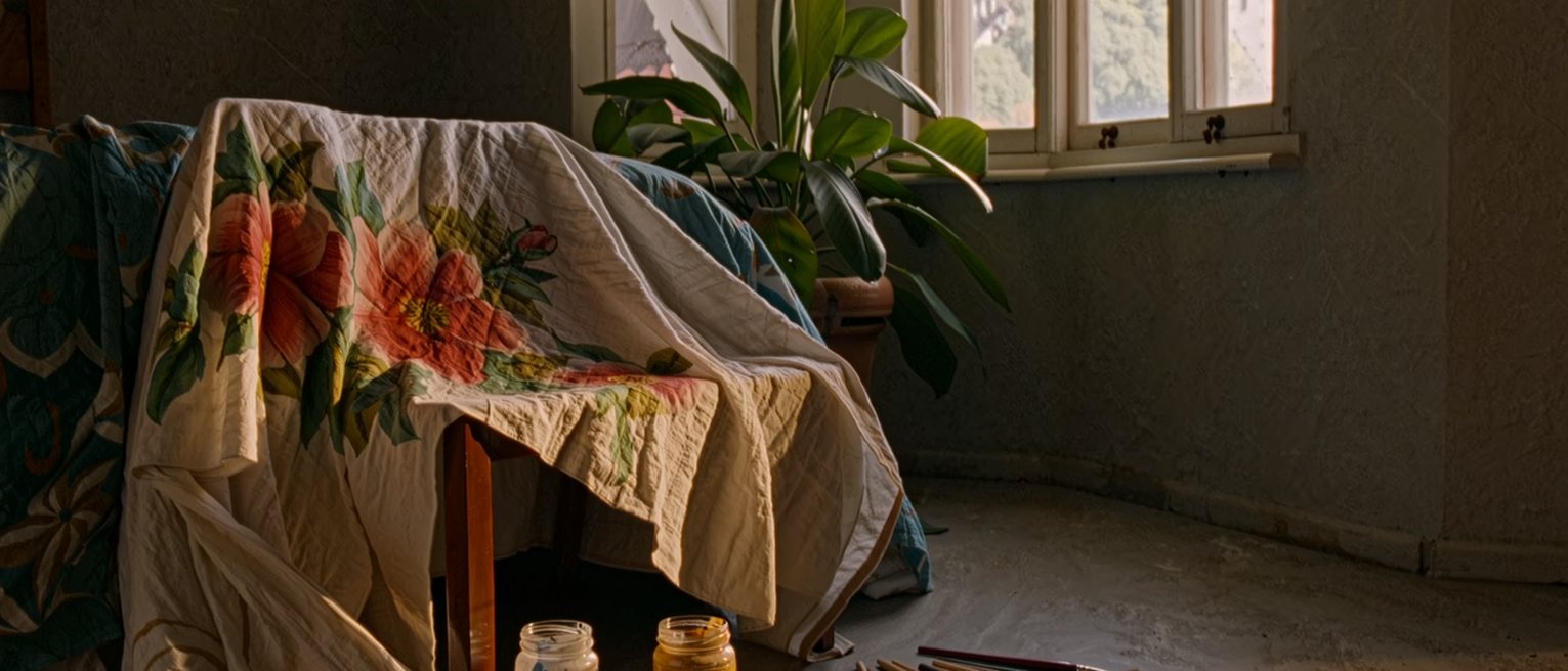 a bedroom seen close with a bedsheet painted with flowers and leaves left to dry over the chair, jars of paint and brushes on the floor and a plant on the sill, the window close behind