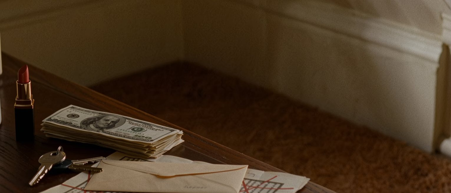 a dressing table seen close with an envelope of cash squared beside a lipstick and a set of house keys, a printed page with its lines crossed through, the lamp soft behind