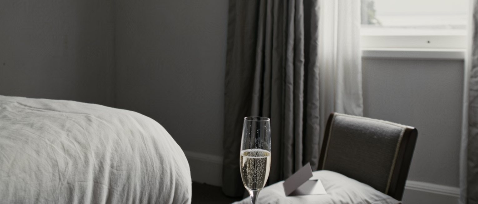 a bedroom seen close with a single glass of champagne on a tray, a chair set beside the bed at a chosen angle and a folded card face down on the pillow, the curtains soft behind