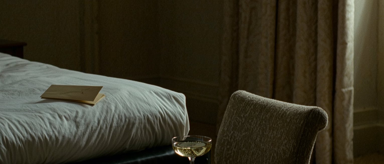 a bedroom seen close with a single glass of champagne on a tray, a chair set beside the bed at a chosen angle and a folded card face down on the pillow, the curtains soft behind
