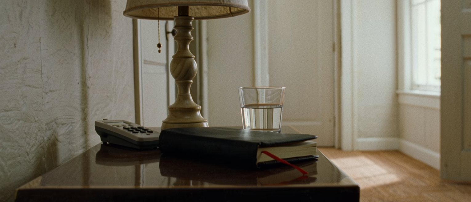 a bedside table seen close with a phone face up beside a diary held shut with an elastic band, a glass of water and the lamp close behind