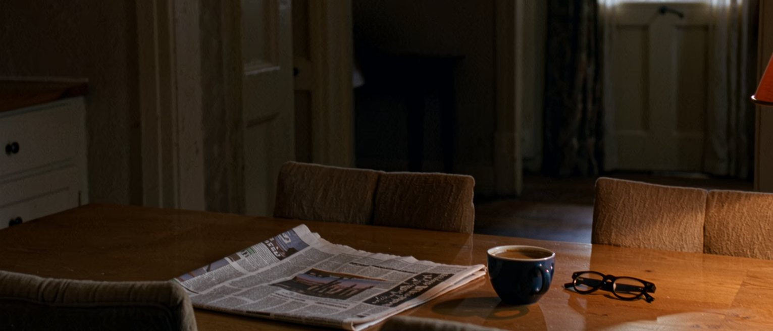 a kitchen table seen close with a local newspaper folded open, a cup of coffee and a pair of reading glasses, three chairs around it and the window soft behind