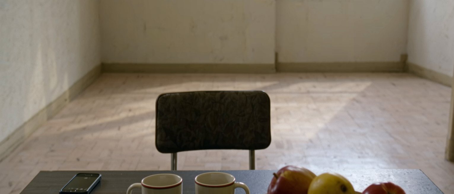 a small flat's table seen close, a phone left beside a note folded once, two cups and a bowl of fruit, the chair opposite pushed in square behind them