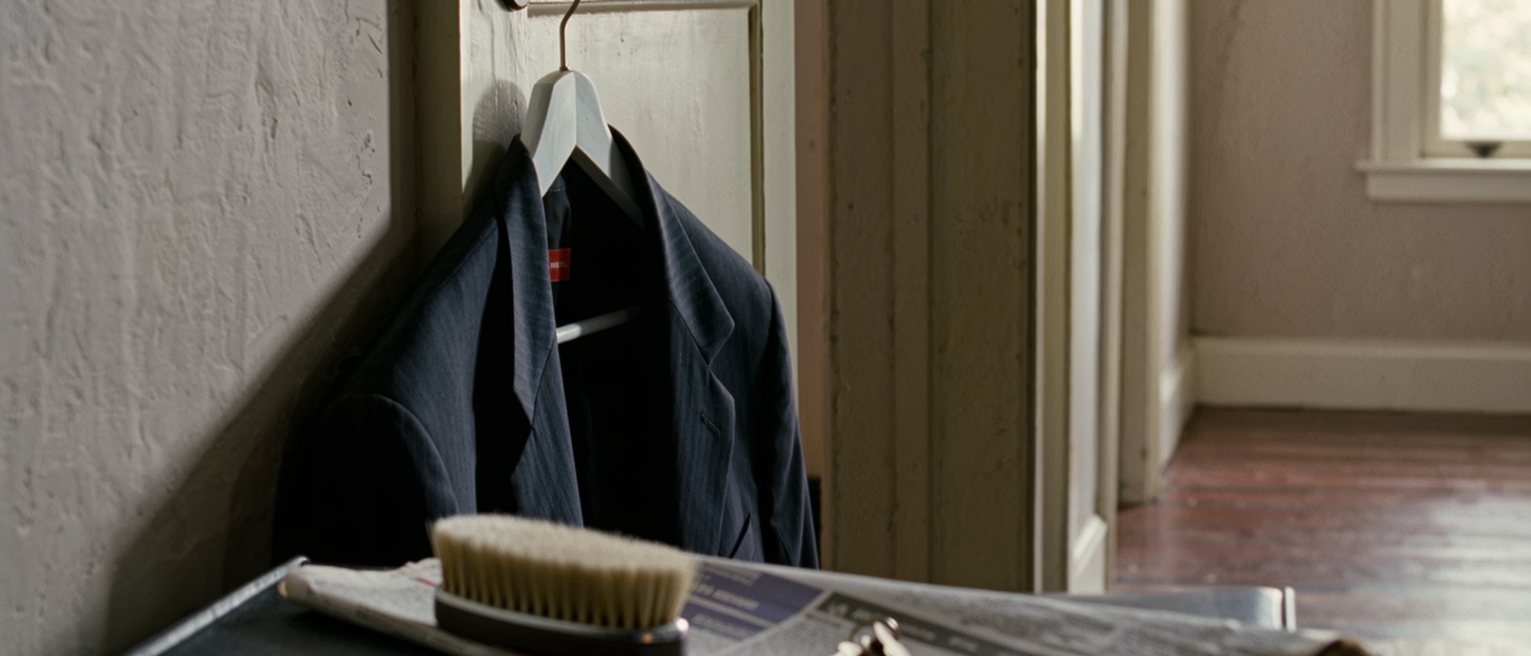 a hall seen close with a suit on its hanger brushed and ready beside a briefcase with nothing in it, a folded newspaper of job listings and a set of keys, the door soft behind