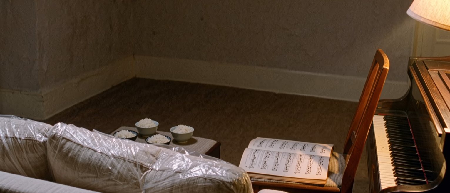 a sitting room seen close with a new sofa still in its plastic, a piano book on the chair and four bowls of rice on the low table, the window soft behind