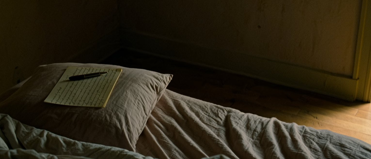 a bedroom with the covers turned down and a written list left on the pillow, the pen still on it, most of the page blank, the headboard and a lit bedside lamp close behind