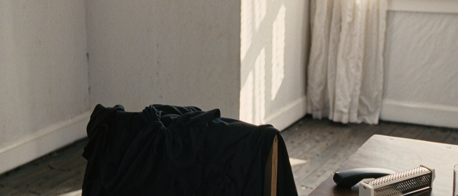 a dressing table seen close with a black dress laid over the chair, a phone face down beside a cheese grater still in its packaging, a card in its envelope and a glass of water, the curtain soft behind