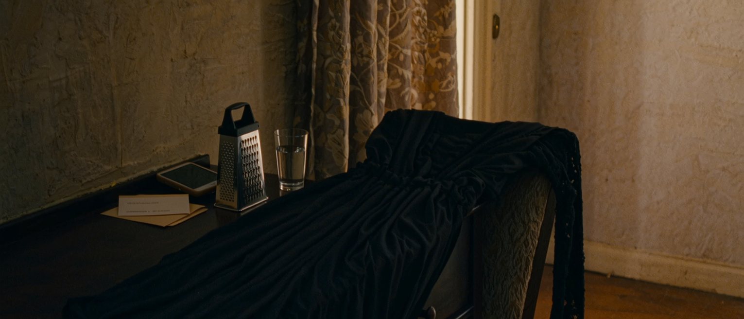 a dressing table seen close with a black dress laid over the chair, a phone face down beside a cheese grater still in its packaging, a card in its envelope and a glass of water, the curtain soft behind