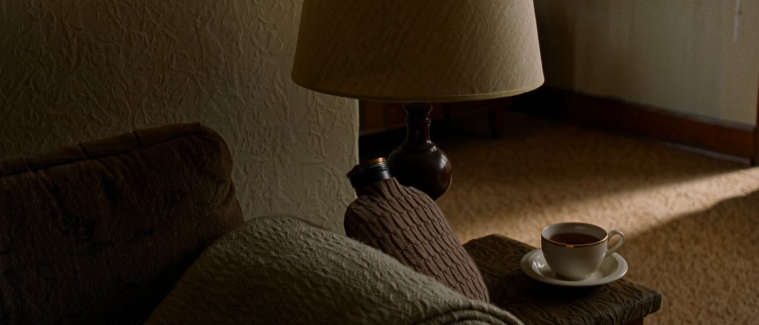 a sofa seen close with a folded wool blanket on one arm, a hot-water bottle in a knitted cover and a cup of tea on the low table, the lamp close behind