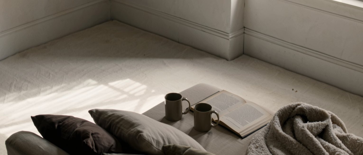 a sofa heaped with cushions and a blanket pushed to one end, two mugs and an open book on the low table in front of it, a Tuesday's worth of nothing happening