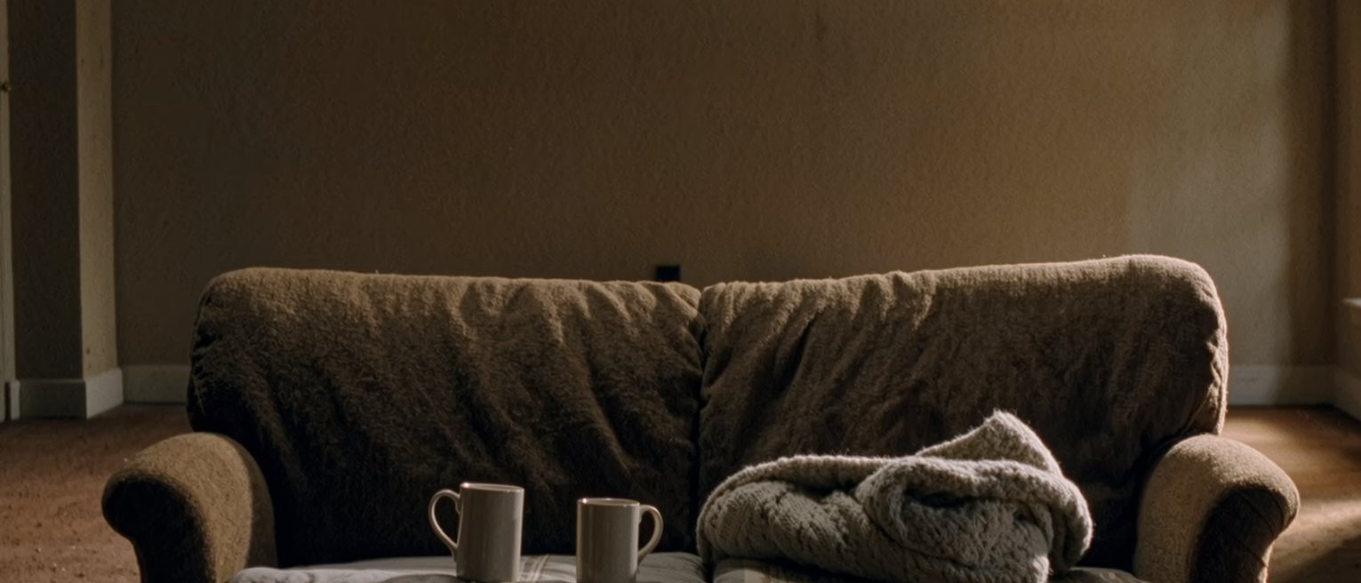 a sofa heaped with cushions and a blanket pushed to one end, two mugs and an open book on the low table in front of it, a Tuesday's worth of nothing happening