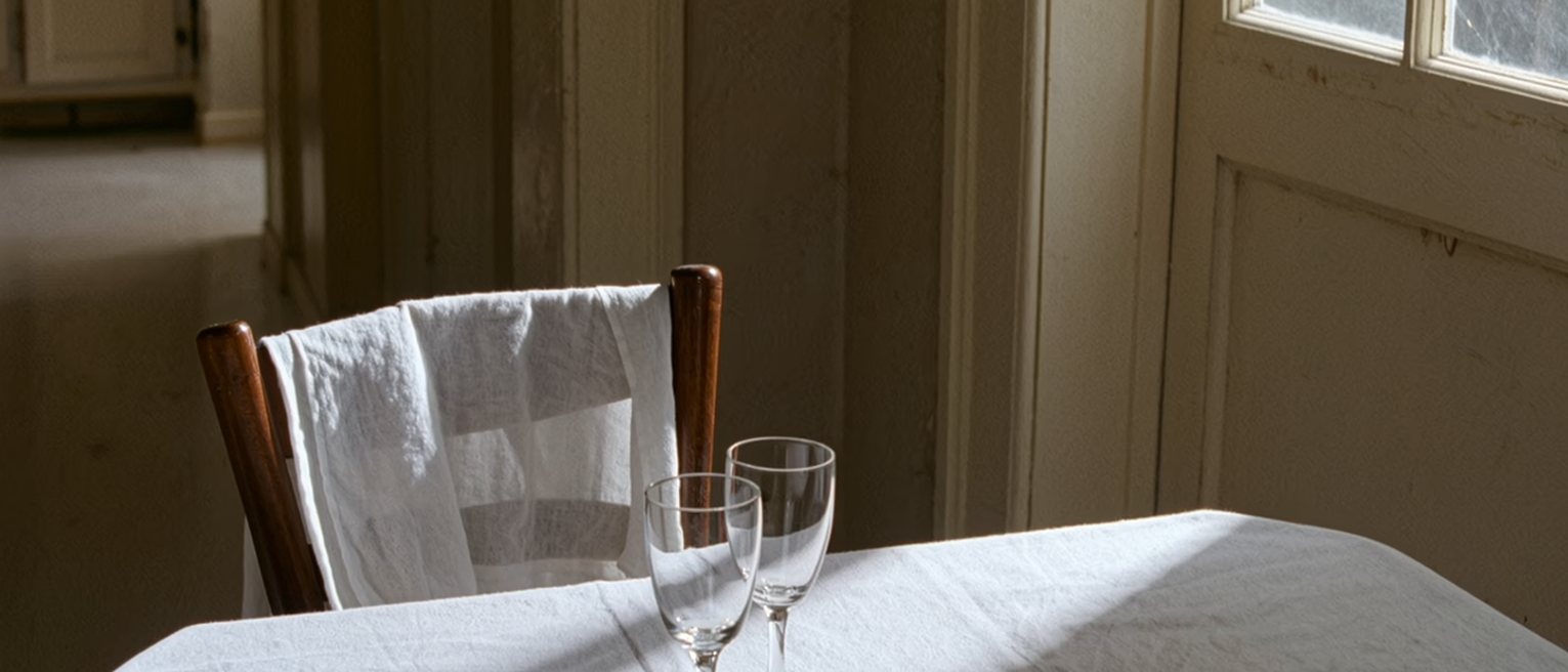 a kitchen with a single place set at the table as it has been set for a long time, the second chair pushed under and a cloth over the good glasses