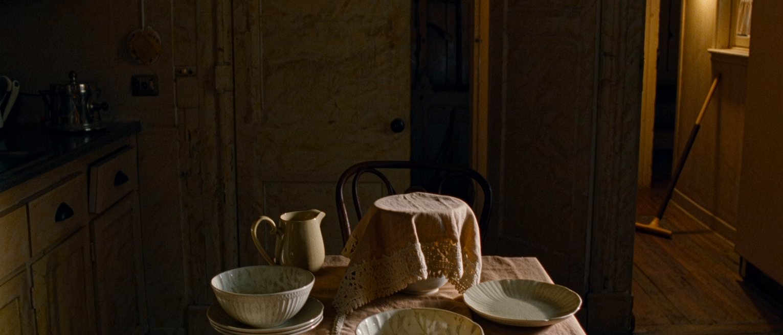 a kitchen with a single place set at the table as it has been set for a long time, the second chair pushed under and a cloth over the good glasses