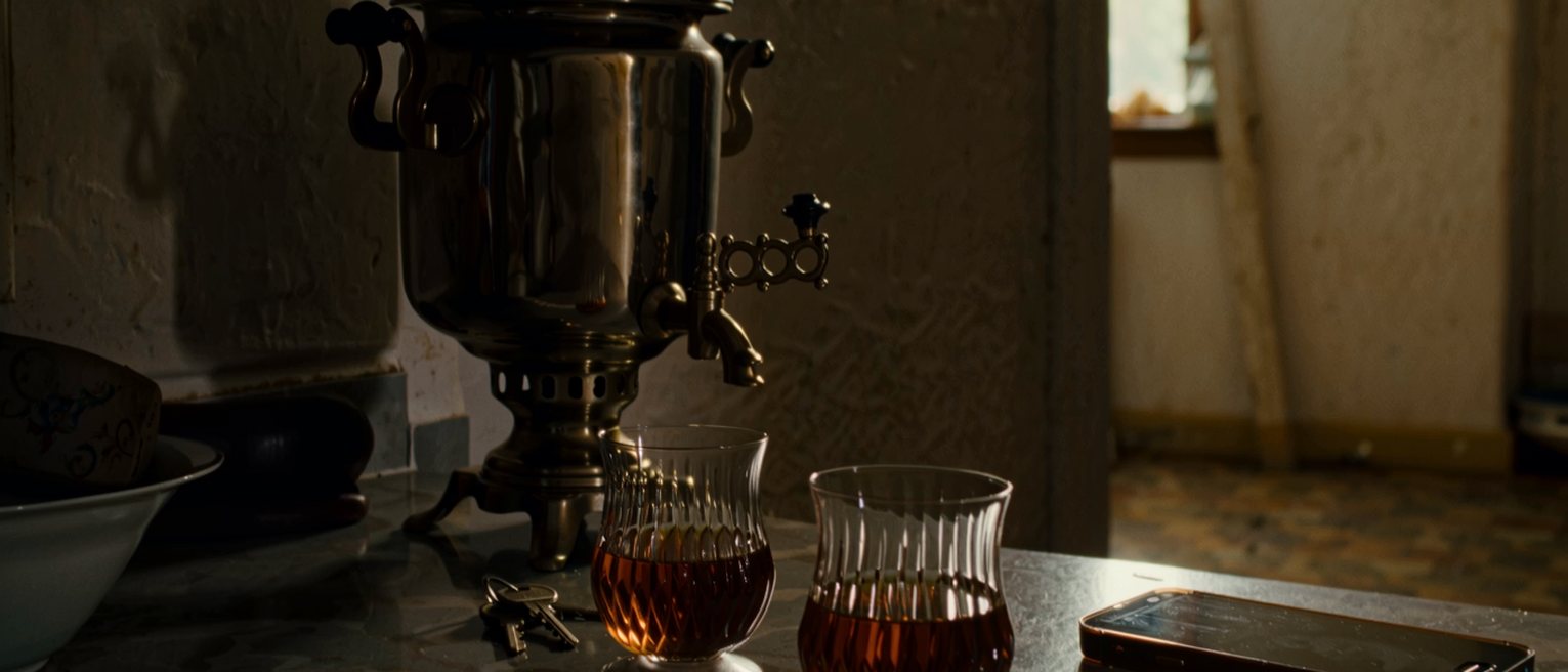 a kitchen table seen close with two tulip tea glasses, a folded newspaper and a phone face down, a set of keys and the samovar soft behind